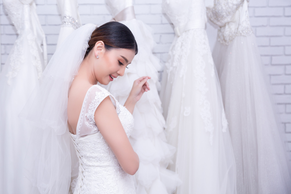A woman in a white wedding dress and veil stands in front of a row of white bridal gowns on mannequins against a white brick wall.