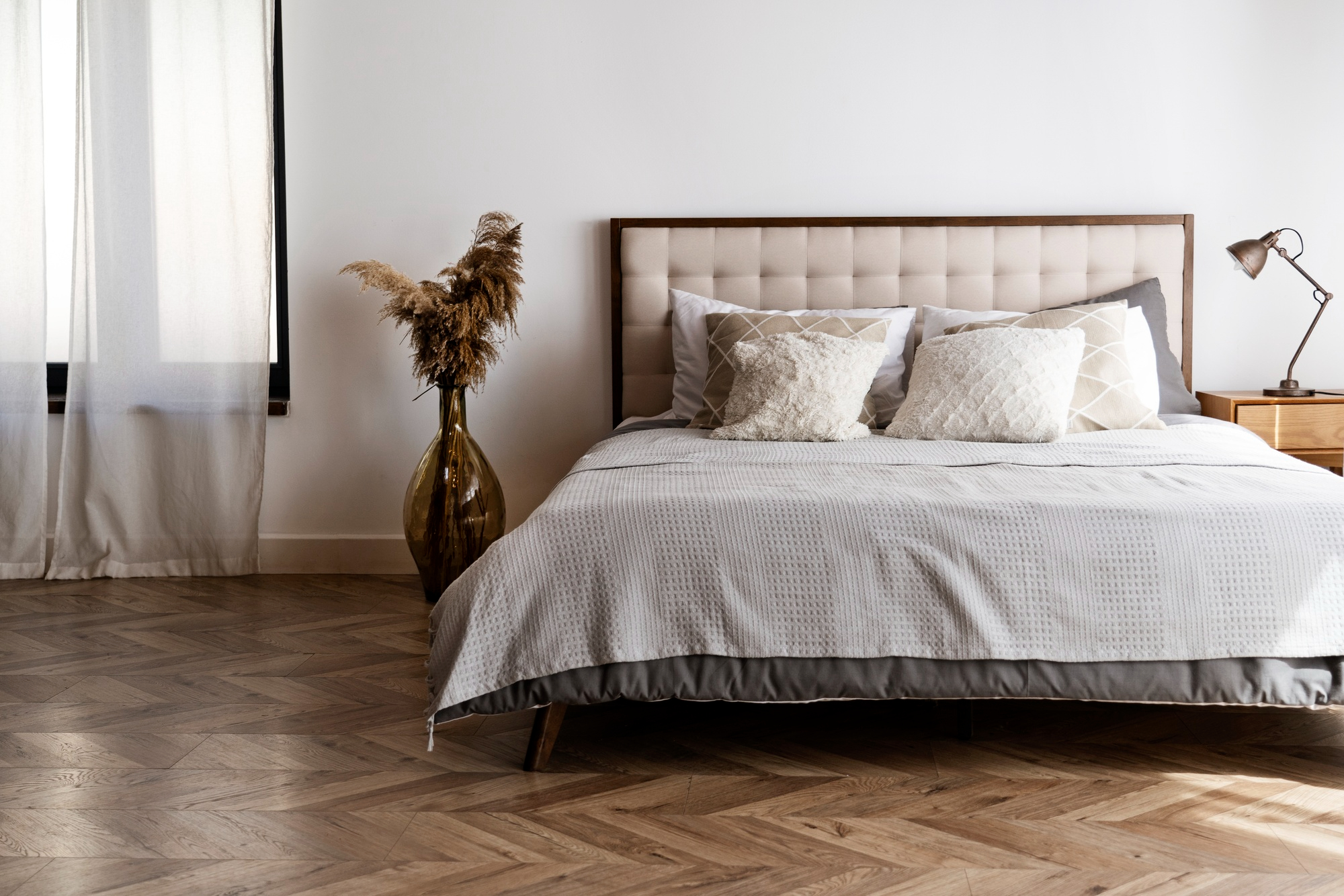 A modern bedroom with a beige upholstered bed, neutral bedding, decorative pillows, a wooden nightstand with a lamp, and a large vase with dried plants on a parquet floor.