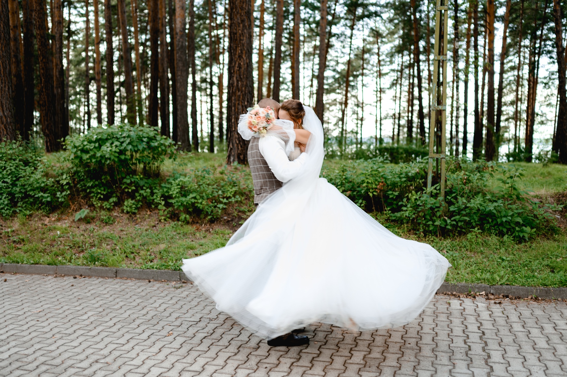 A bride and groom embrace and twirl on a paved path outdoors, surrounded by greenery and trees.