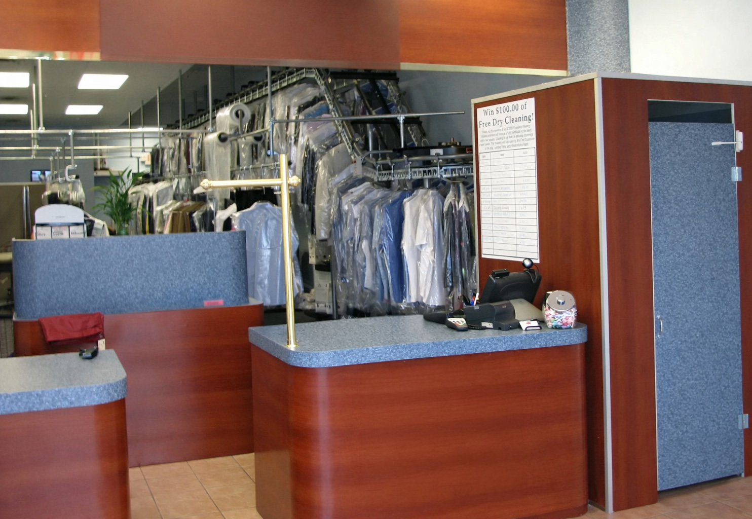 Interior of a dry cleaning store with a counter, cash register, and racks of clothes covered in plastic hanging in the background.
