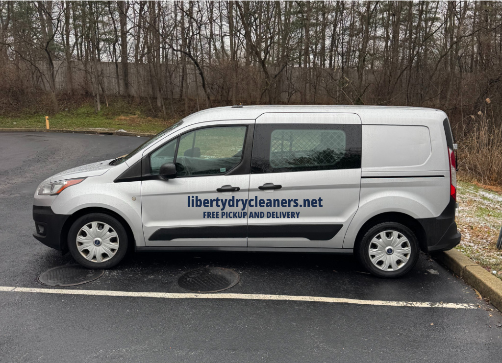 A gray van in a parking lot with "libertydrycleaners.net FREE PICKUP AND DELIVERY" printed on the side; trees are visible in the background.