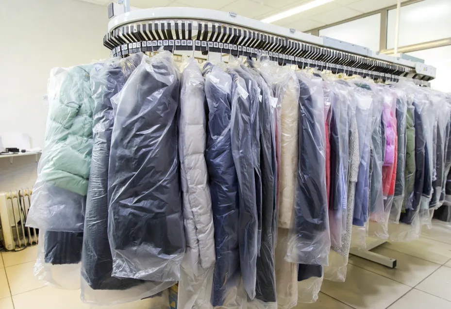 A row of assorted coats and jackets covered in plastic garment bags hangs on a conveyor rack in a dry cleaning facility.