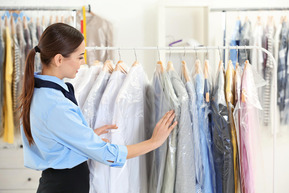 A woman in a blue shirt examines clothes covered in plastic hanging on a garment rack in a dry cleaning shop.