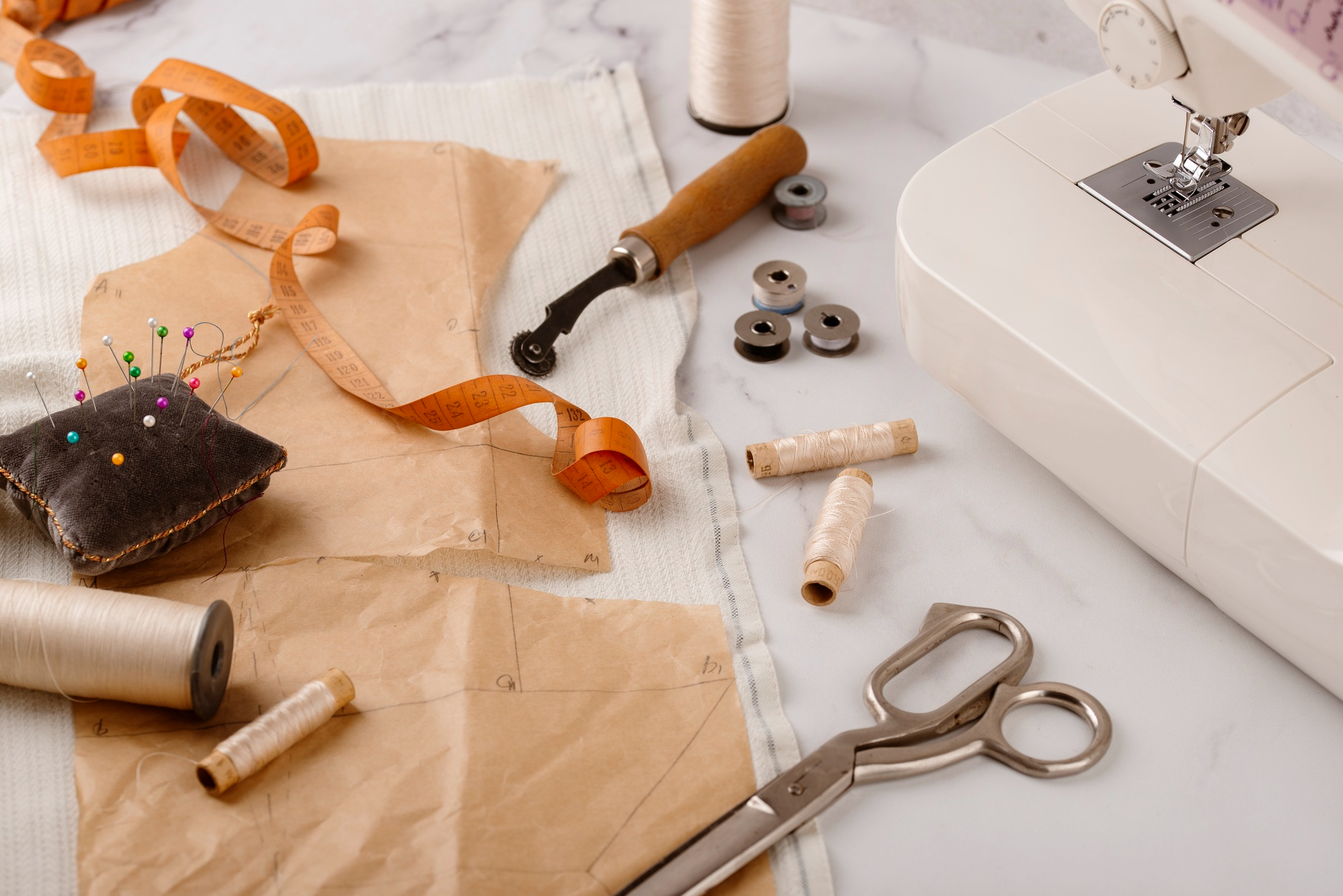 Sewing materials on a table, including paper patterns, a pincushion, measuring tape, scissors, thread spools, bobbins, a tracing wheel, and a sewing machine.
