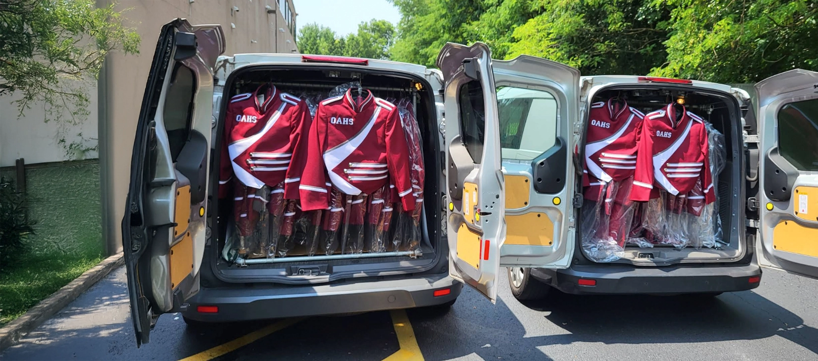 Two open vans parked side by side, each filled with rows of red and white "OARS" uniforms on hangers in garment bags, parked on an asphalt surface beside a building and trees.