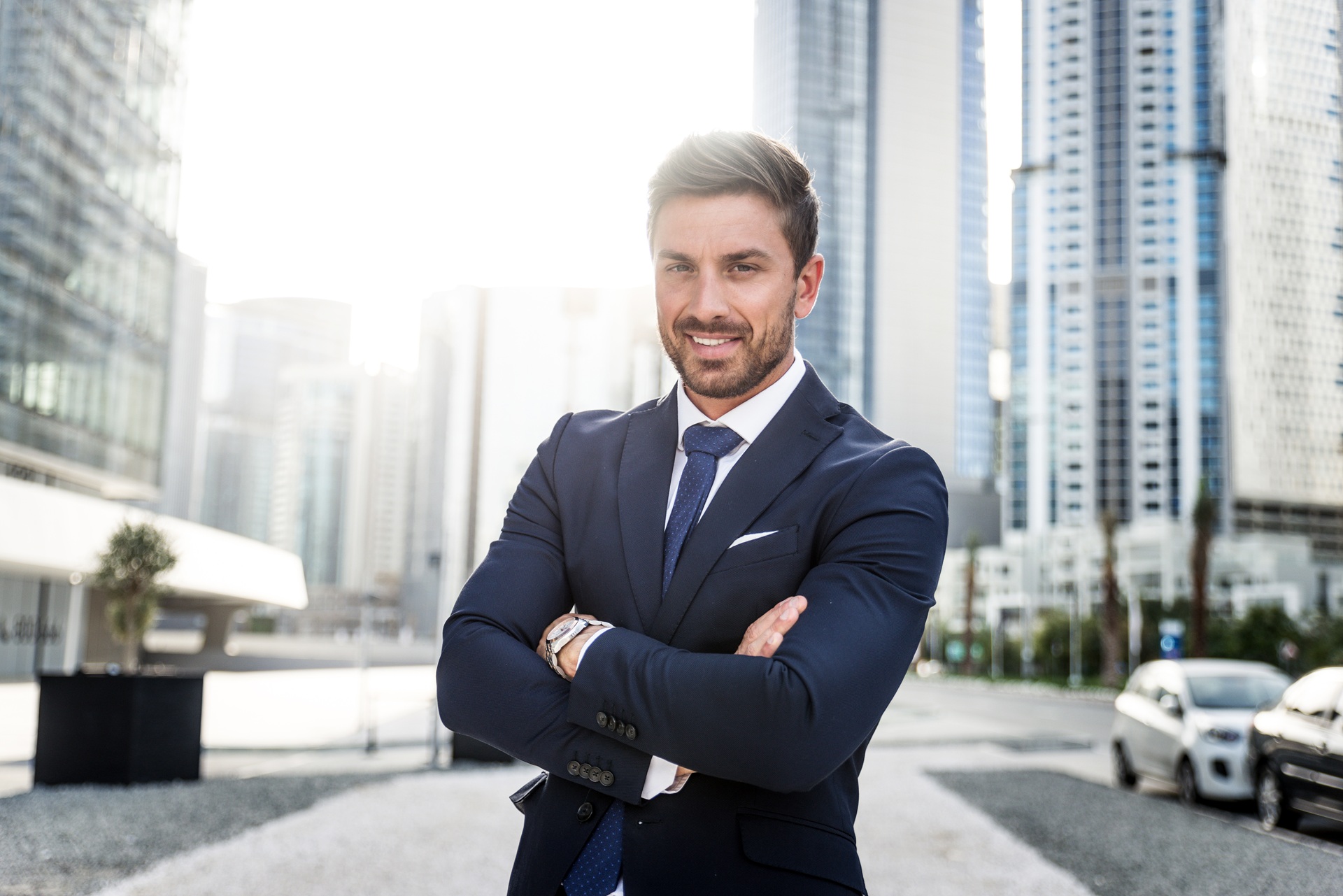 A man in a suit stands outdoors with arms crossed, smiling, in front of tall modern office buildings in a city setting.