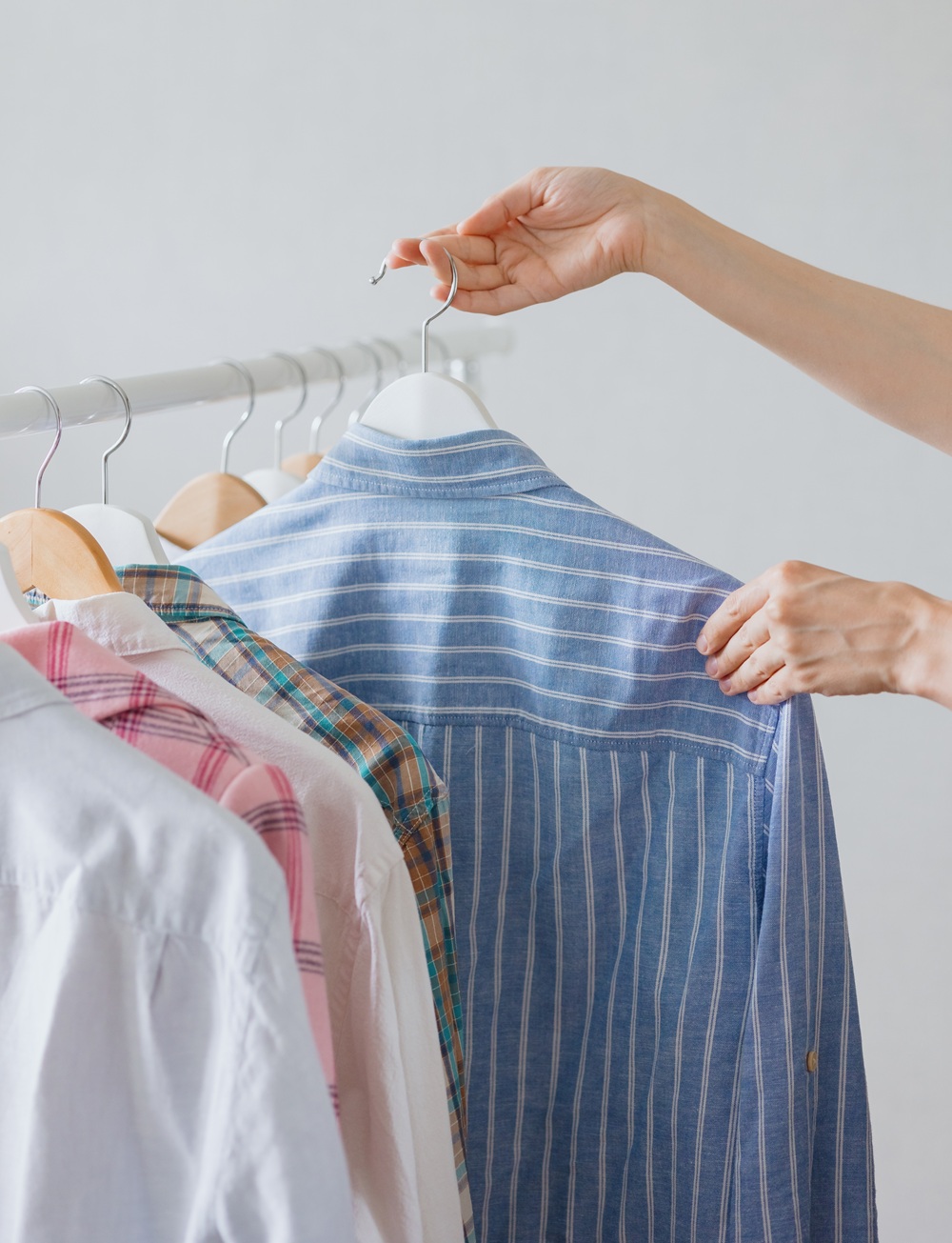 A person selects a blue striped shirt from a row of assorted shirts hanging on wooden and white hangers on a clothing rack.