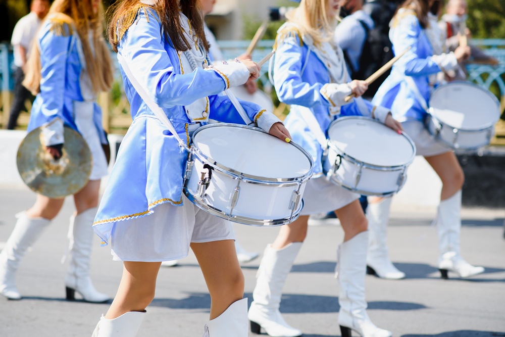 A group of people in matching blue uniforms and white boots march outdoors, playing snare drums and cymbals.