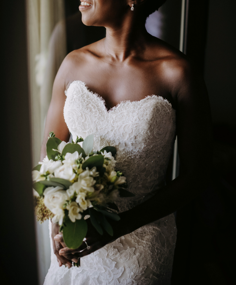A bride in a strapless lace wedding dress holds a bouquet of white flowers and greenery, standing by a window with soft light.