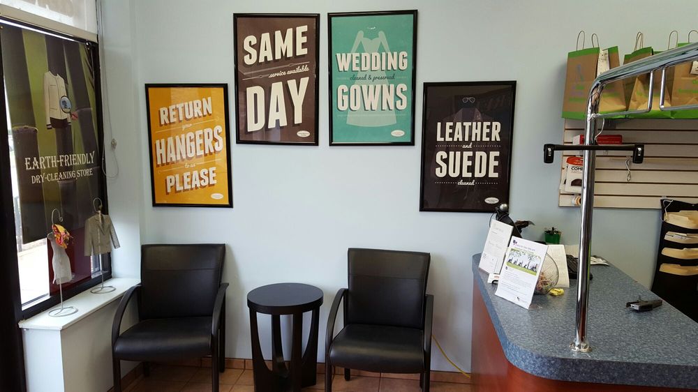 A small waiting area with two black chairs, a round table, wall signs about dry cleaning services, and part of a counter with informational pamphlets.