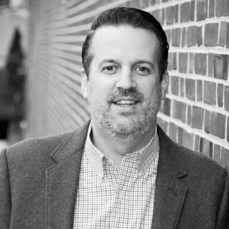 Black and white photo of a man with short dark hair and a beard, wearing a blazer and checkered shirt, standing against a brick wall.