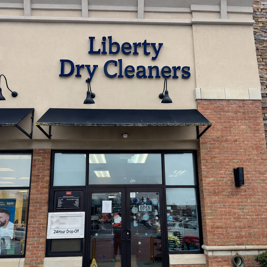 Storefront of Liberty Dry Cleaners with large blue sign, glass door displaying an "Open" sign, and a 24-hour drop-off notice posted on the left window.