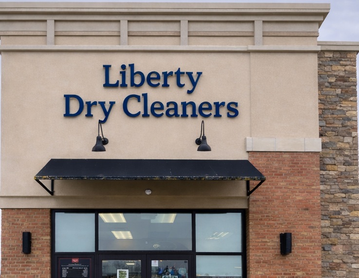The exterior of Liberty Dry Cleaners with a glass entrance, stone and brick facade, and a black awning above the door.
