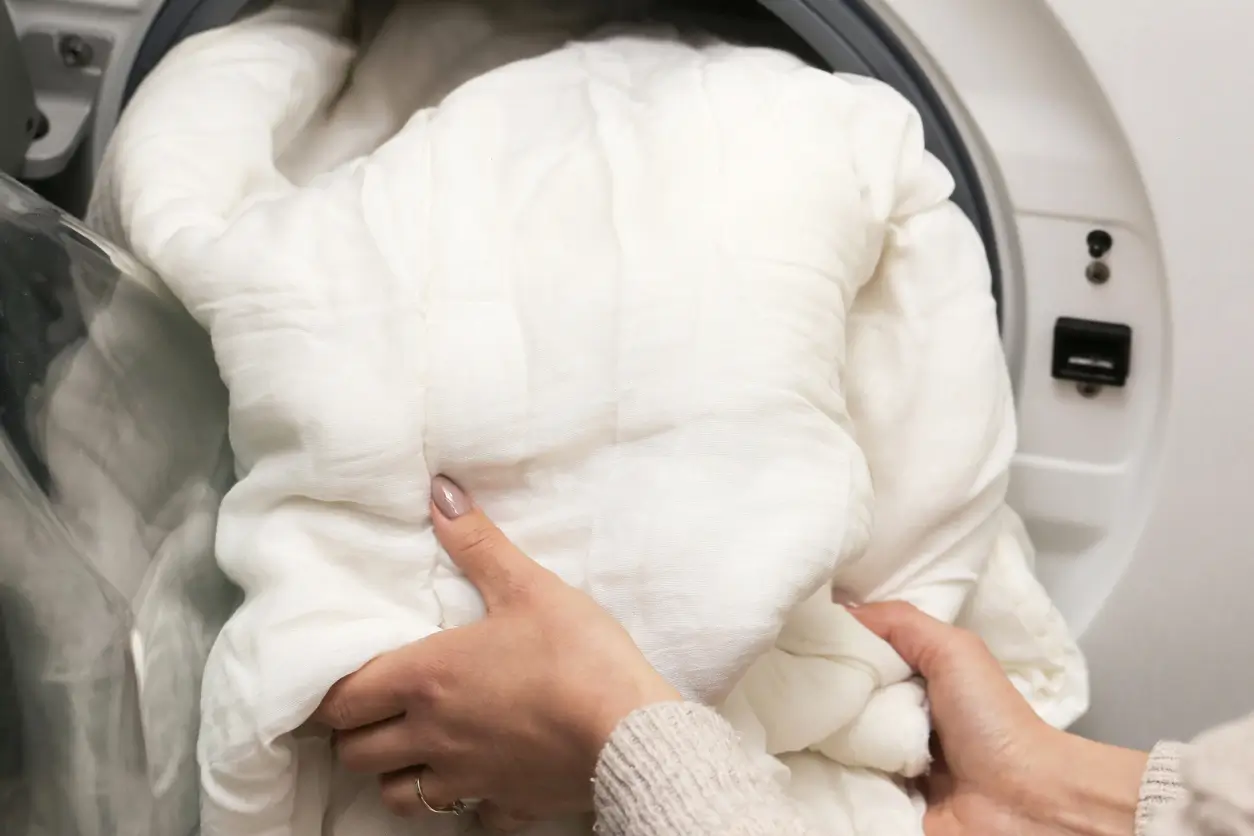 Person placing a large white comforter into the open door of a washing machine.
