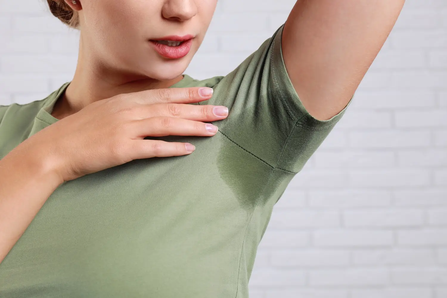 A person in a green shirt with a visible sweat stain under their arm, touching their armpit, against a white brick background.